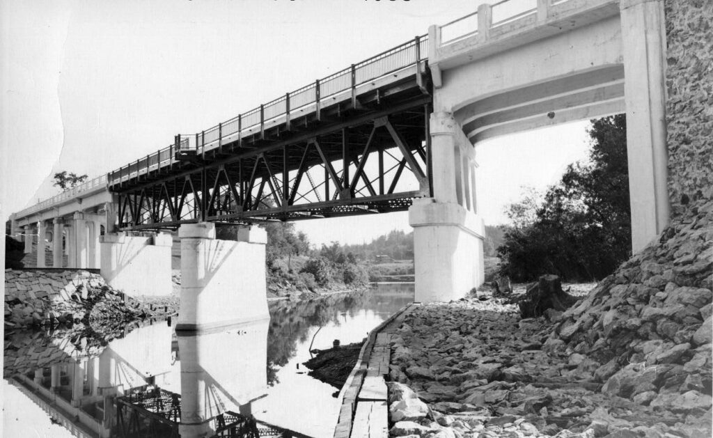 A black-and-white image of a bridge with a metal truss and concrete supports spans a river. Rocky shoreline and vegetation are visible below the bridge.