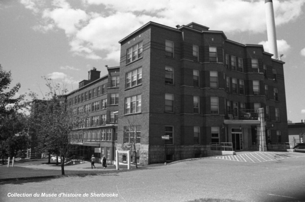 Black and white photo of a large, multi-story brick building with many windows. A few people walk near the entrance. The left corner of the image shows trees, and there's a chimney on the right side.