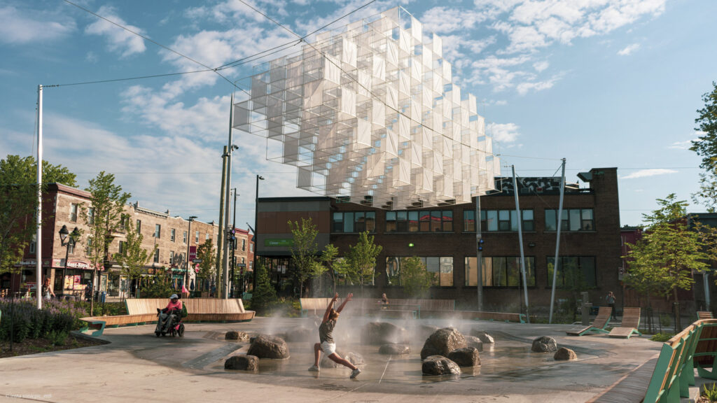 Person joyfully running through a modern urban fountain installation with geometric art overhead, surrounded by buildings and trees under a sunny sky.