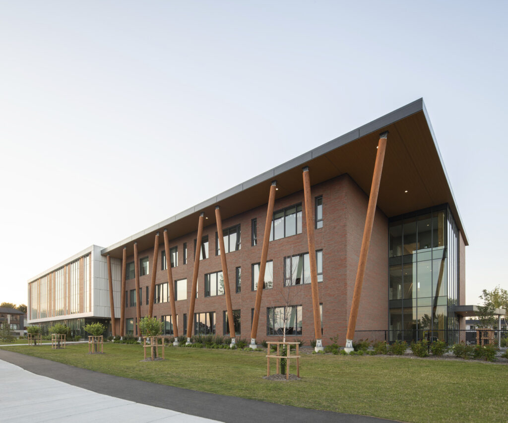 A modern, multi-story building with large, angled wooden beams, featuring a mix of glass and brick exterior, situated on a landscaped area with a walkway.