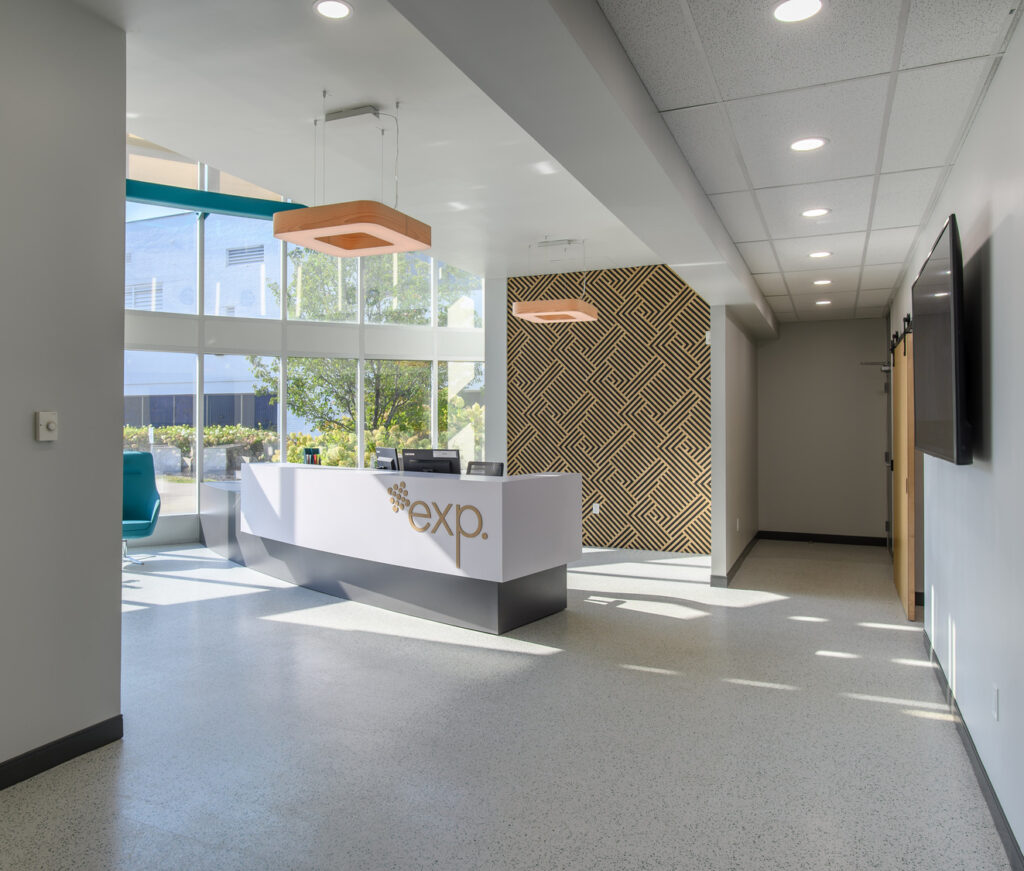 A modern office reception area with a white reception desk labeled "exp," a patterned feature wall, large windows, and clean, minimalist decor.