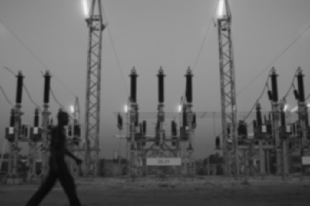 A person walks past electrical equipment at a power substation. Tall structures with wires and insulators are prominently visible. The scene is in grayscale.