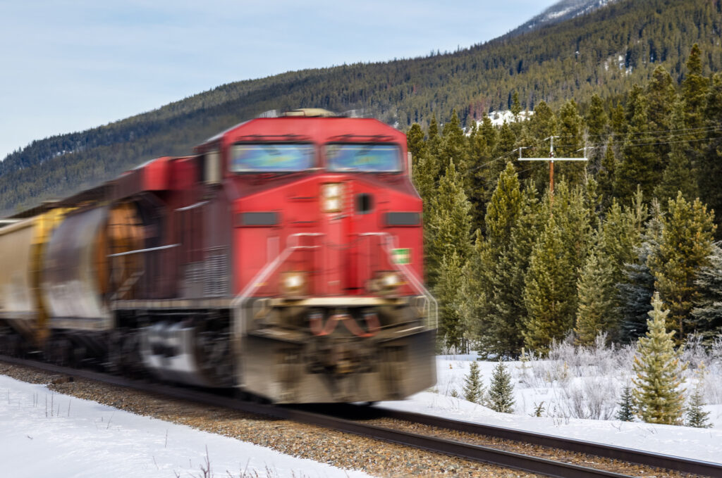 A red freight train moves at high speed on a snowy track through a mountainous forest landscape.