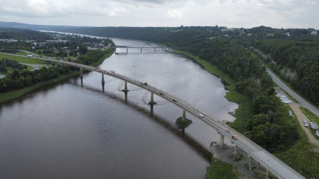 Aerial view of a large river with two bridges spanning it; one bridge is closer with vehicles on it and appears under construction, while the other is further away. Forested areas and roads flank the river.