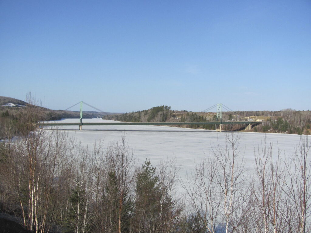 A large river in a cold region with two green suspension bridges in the background. The river is largely frozen with sparse trees in the foreground. The sky is clear and blue.
