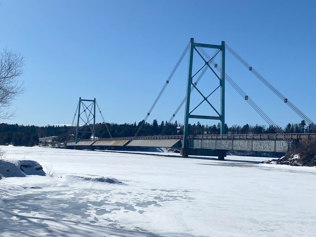 A green suspension bridge spans over a frozen, snow-covered river on a clear, sunny day with a forested backdrop.