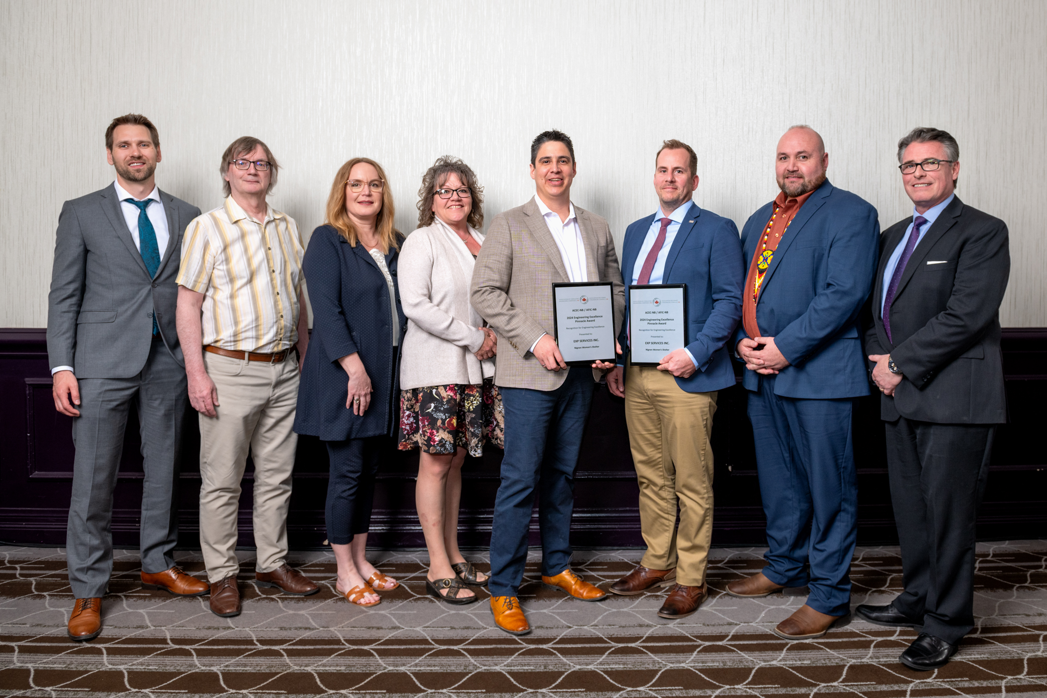A group of eight people standing in a row, with two individuals holding framed certificates. They are dressed in business or casual attire and posing against a plain wall.