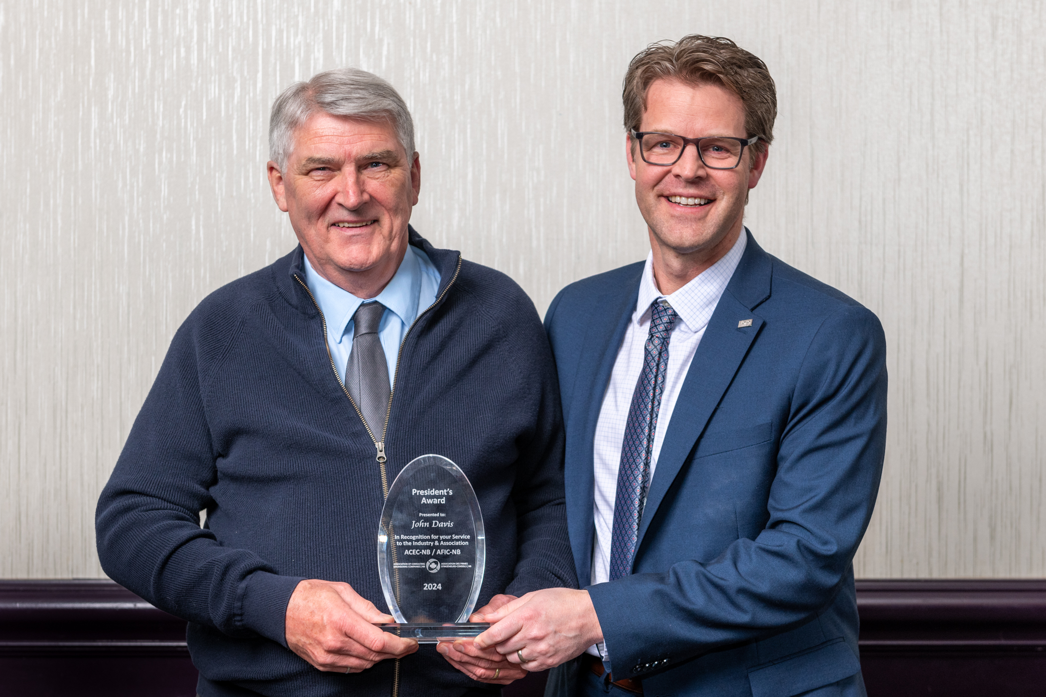 Two men standing side-by-side holding an award. The man on the left is wearing a blue sweater and the man on the right is wearing a blue suit and tie. They are both smiling at the camera.