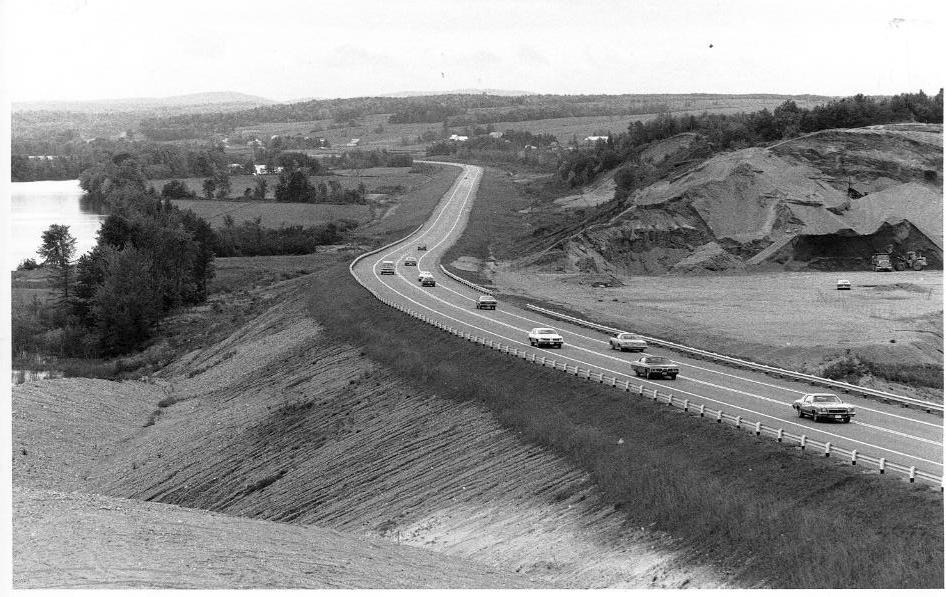 A black and white photo of a rural highway with several cars driving in both directions, flanked by trees and a body of water on one side and a hillside on the other.