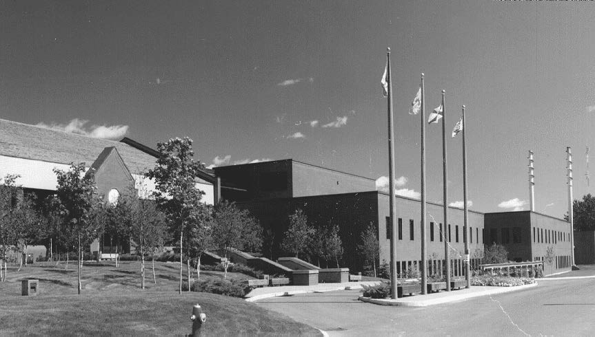 Black and white photo of a large, modern brick building with a clock, surrounded by trees and flagpoles with flags. Driveway and fire hydrant are visible in front.