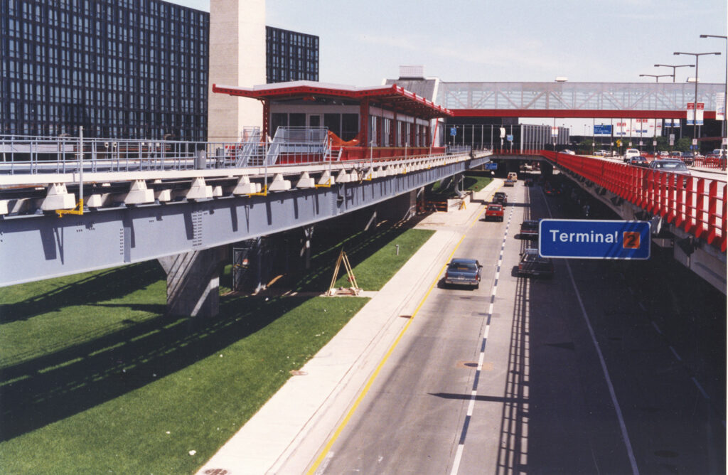 View of an airport terminal roadway with a suspended monorail track and station to the left. Blue sign indicates directions to Terminal 2. Cars are driving along the road.
