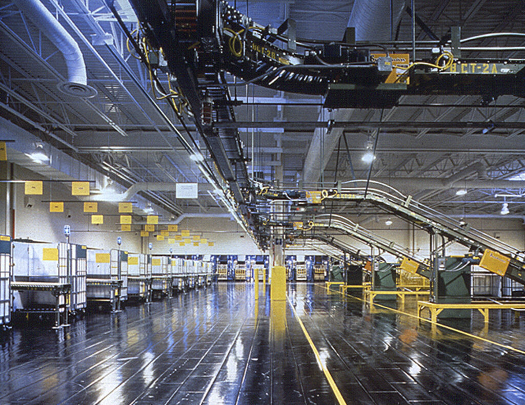 An empty, well-lit industrial facility with conveyor belts, machinery, and storage areas. The metallic floor is reflective, and the ceiling has exposed pipes and ducts.