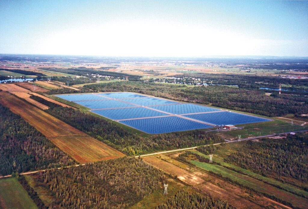 Aerial view of a large solar farm with numerous solar panels in a rural area surrounded by trees, fields, and distant towns.
