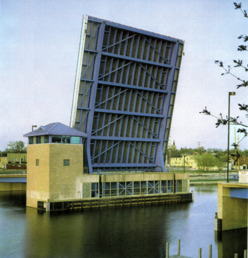 A blue drawbridge in the raised position spans over a body of water, with trees and buildings visible in the background.