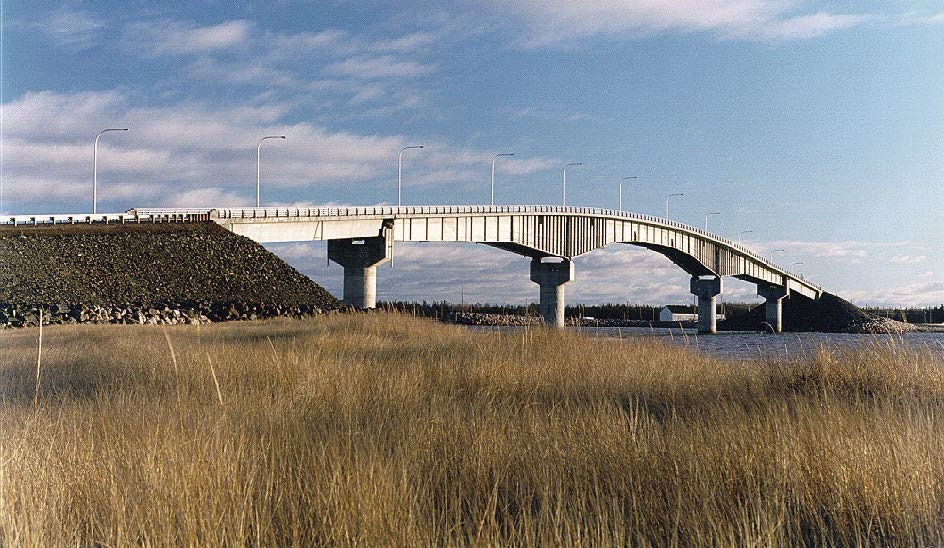 A long, slightly arched bridge extends over a body of water, supported by concrete pillars, with tall grass in the foreground and a partly cloudy sky.