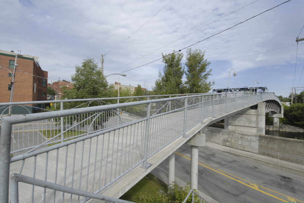 A concrete pedestrian bridge with metal railings extends over a street and connects to a larger road bridge under a partly cloudy sky. Buildings and trees are visible in the background.