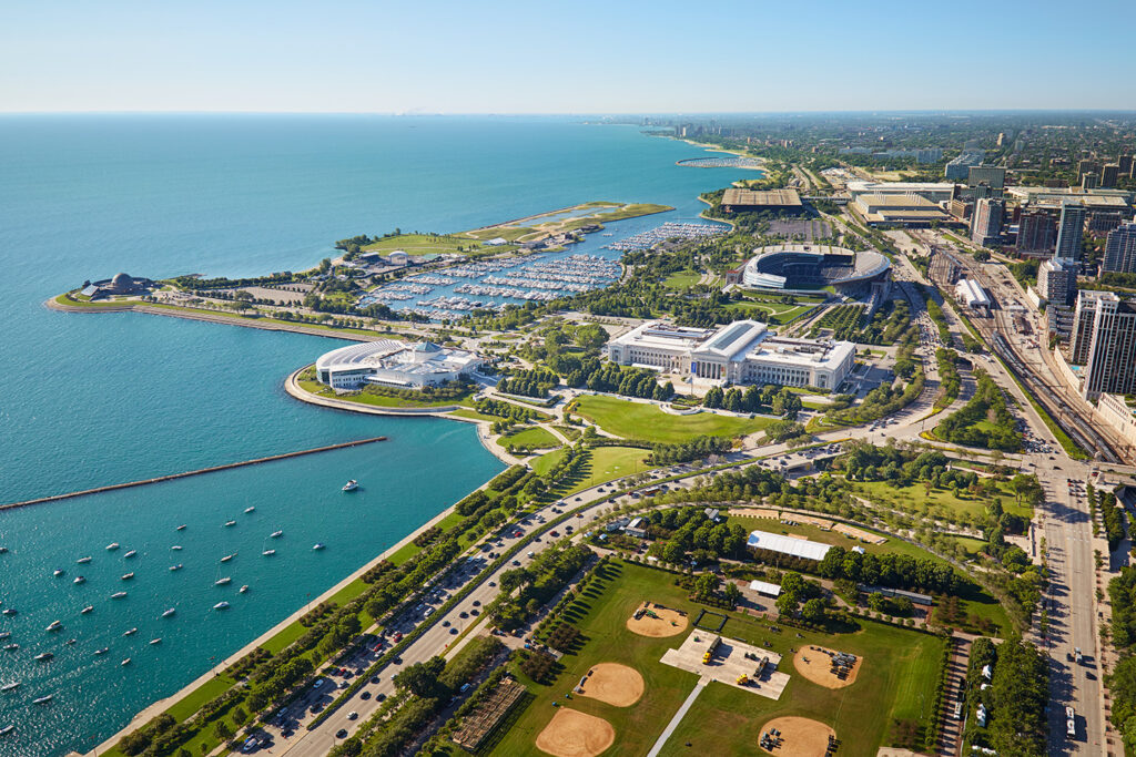 Aerial view of a lakefront city with parks, beaches, sports fields, and buildings, including a stadium near the shoreline. Boats are visible in the water, and roads run through the city.