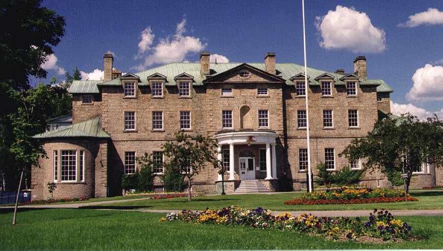 A large stone building with a symmetrical facade, green roof, and multiple windows, surrounded by well-manicured lawns and flower beds, under a blue sky with scattered clouds.