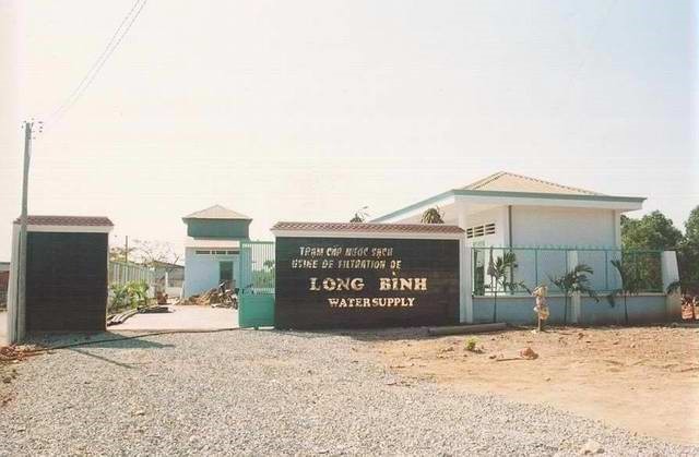 Entrance of a water supply facility with a sign that reads "Long Binh Water Supply." The area is surrounded by a fence and the ground is covered with gravel.