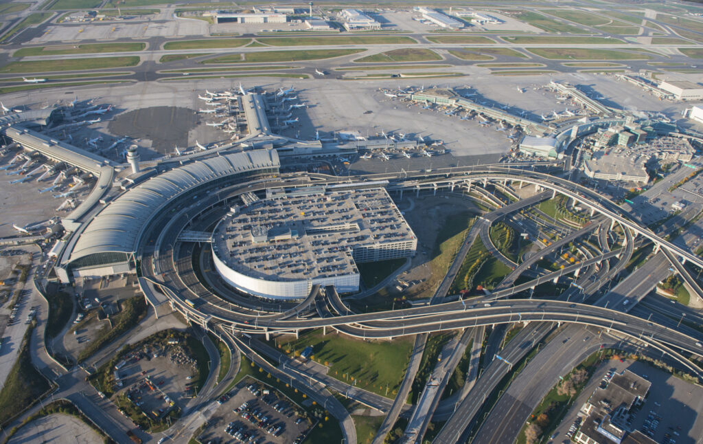 Aerial view of a large international airport with multiple terminals, runways, and parked airplanes. Numerous roads and parking structures surround the terminals.