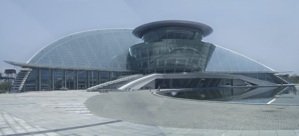 A modern glass and steel building with a distinctive curved design, featuring a large cylindrical structure at the center, surrounded by a spacious paved area and a reflecting pool.
