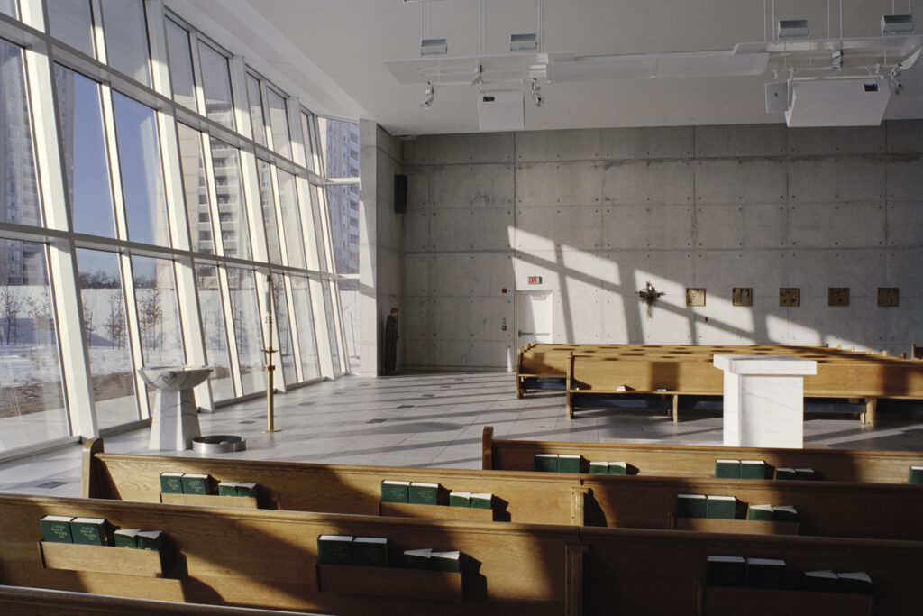 Modern chapel interior with tall windows allowing sunlight to stream in. Pews with hymn books are neatly arranged facing a marble altar. A concrete wall with religious icons is visible in the background.