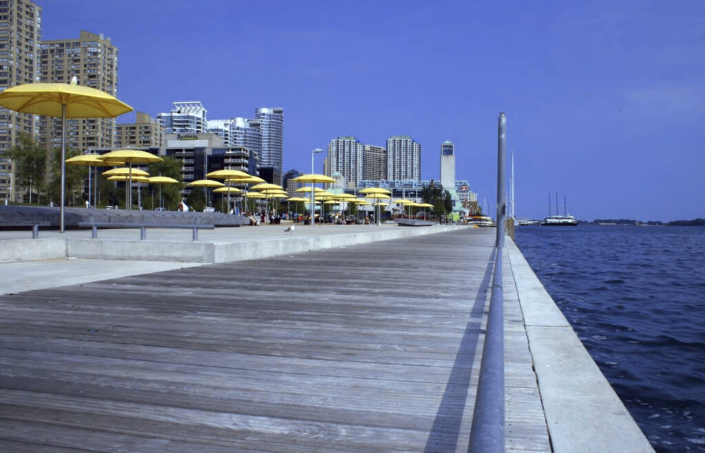 A wooden boardwalk with yellow umbrellas, city buildings, and a body of water on a clear day.