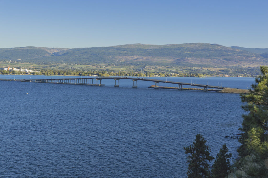 A long, low bridge stretches across a wide body of water with green hills and mountainous terrain in the background under a clear blue sky. Trees are visible in the foreground.