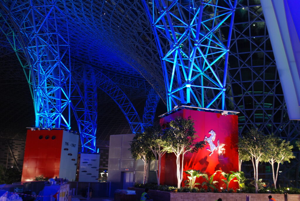 Large indoor structure with intricate blue-lit metal framework and two red walls featuring a silver horse emblem. Several small trees and plants are placed near the base of the red walls.