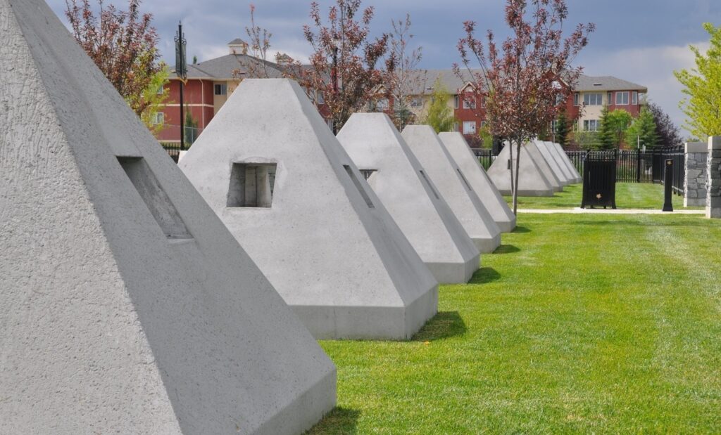 A row of small, gray concrete pyramid structures with square windows, situated on a grassy area, with red buildings and a fence in the background.