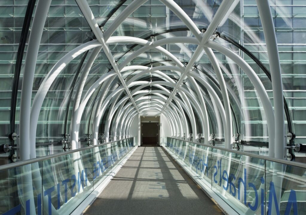 A modern glass and steel pedestrian bridge with an arched design, connecting two buildings.