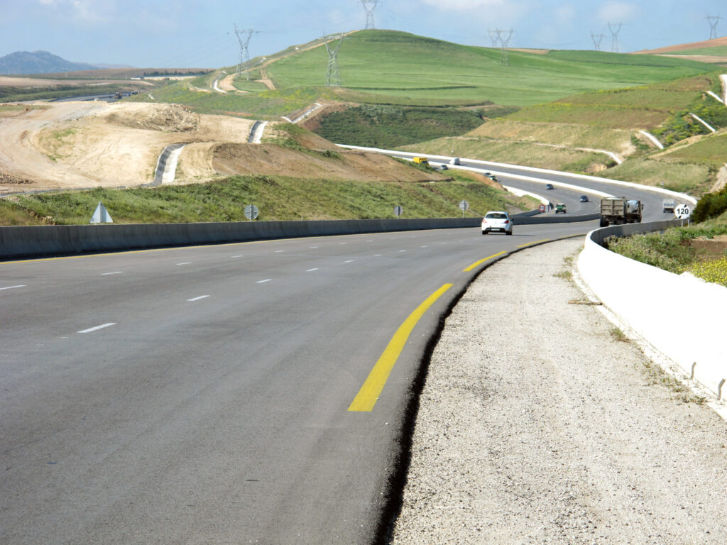 A curved, multi-lane highway with vehicles driving on it, surrounded by rolling green hills and construction areas, with power lines in the background.