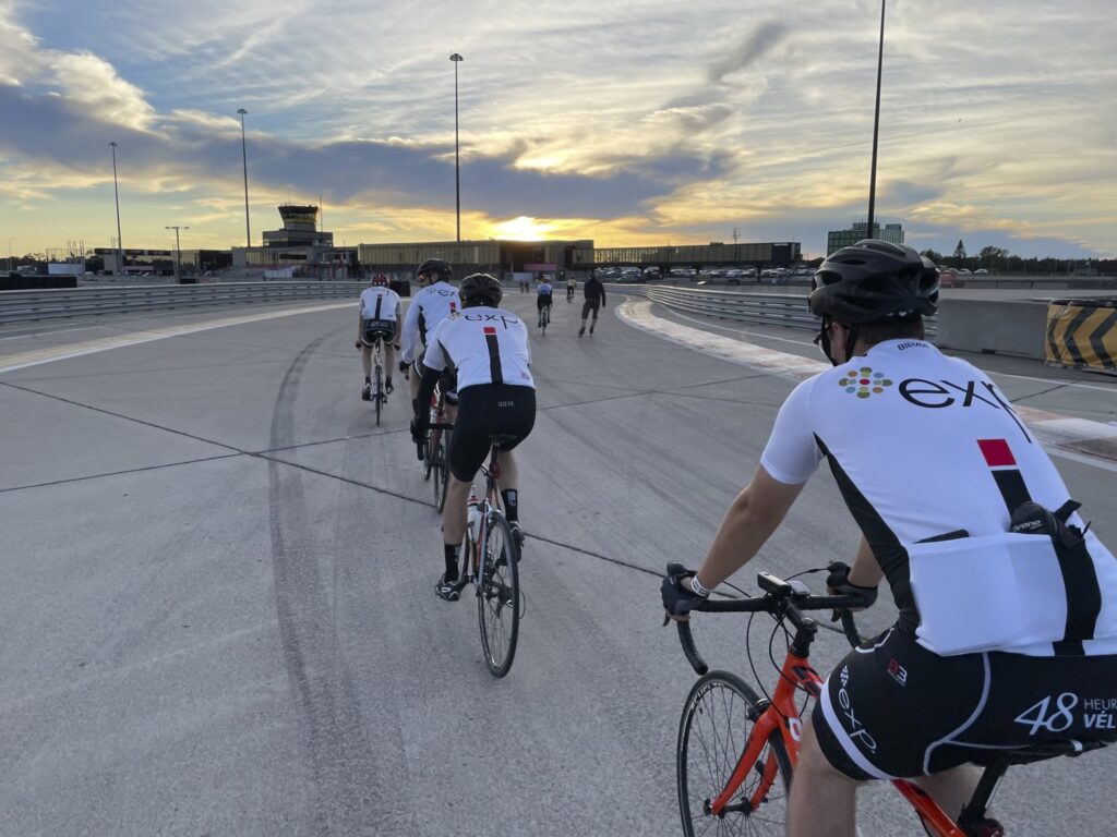 A group of cyclists in branded jerseys ride on an open road at sunset. An airport runway and terminal buildings are visible in the background.