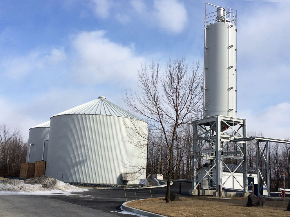 The image shows an industrial facility with two large, white, cylindrical tanks and a tall, silo-like structure standing on a concrete pad. Leafless trees are in the background beneath a partly cloudy sky.
