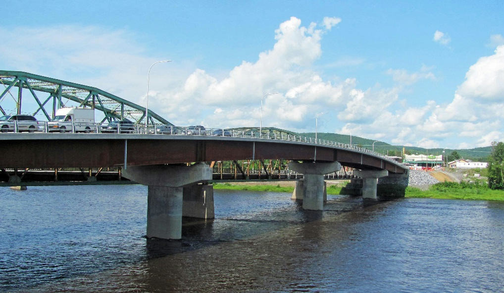 A concrete and steel bridge spans over a body of water with cars visible on the left side. The sky is partly cloudy.