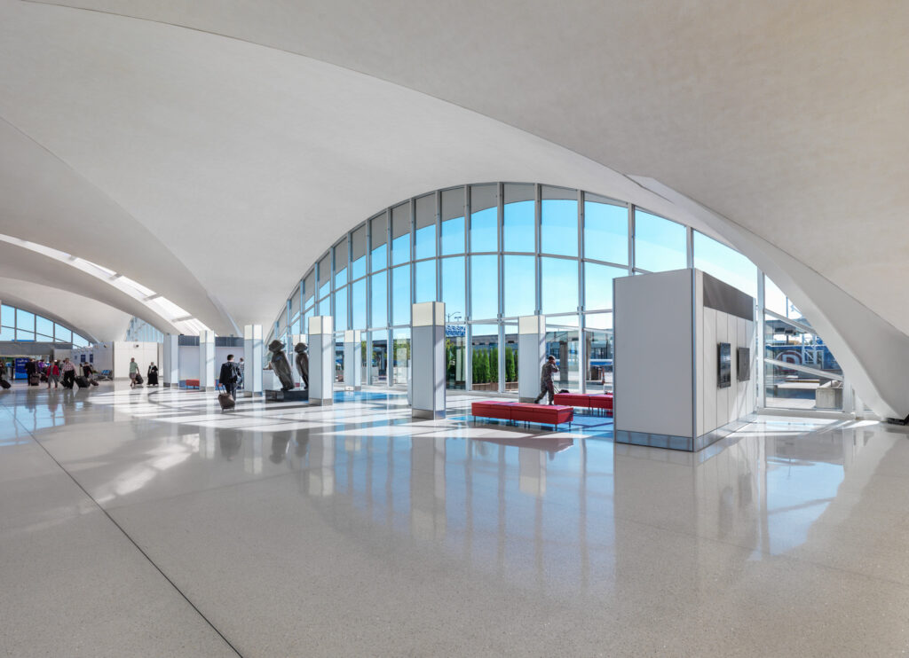 Bright open airport terminal with large arched windows, white interior, shiny floor, and scattered travelers.
