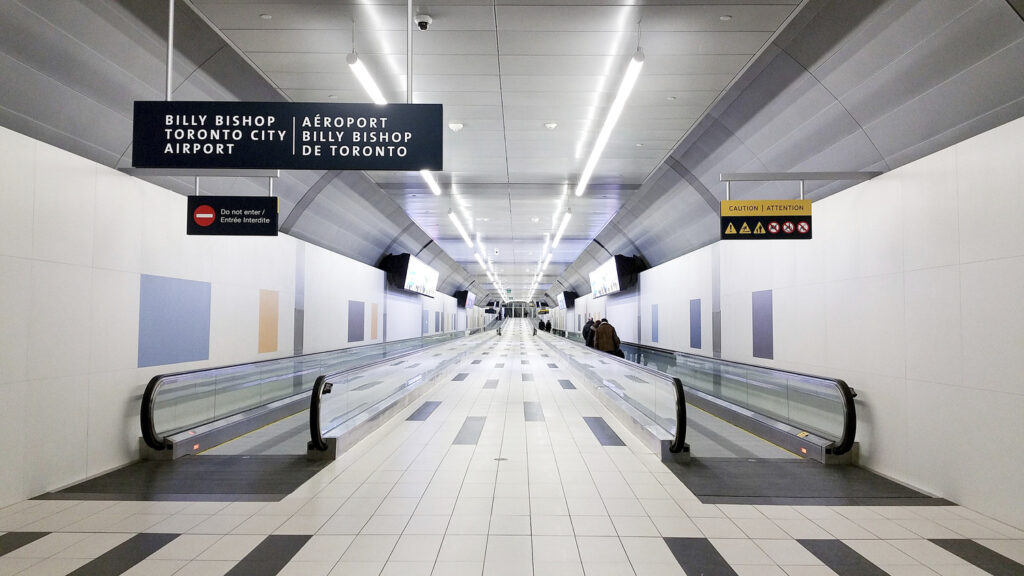 A modern, well-lit airport corridor with moving walkways, displaying signs for Billy Bishop Toronto City Airport. There are safety warnings and directional indicators visible along the walls.