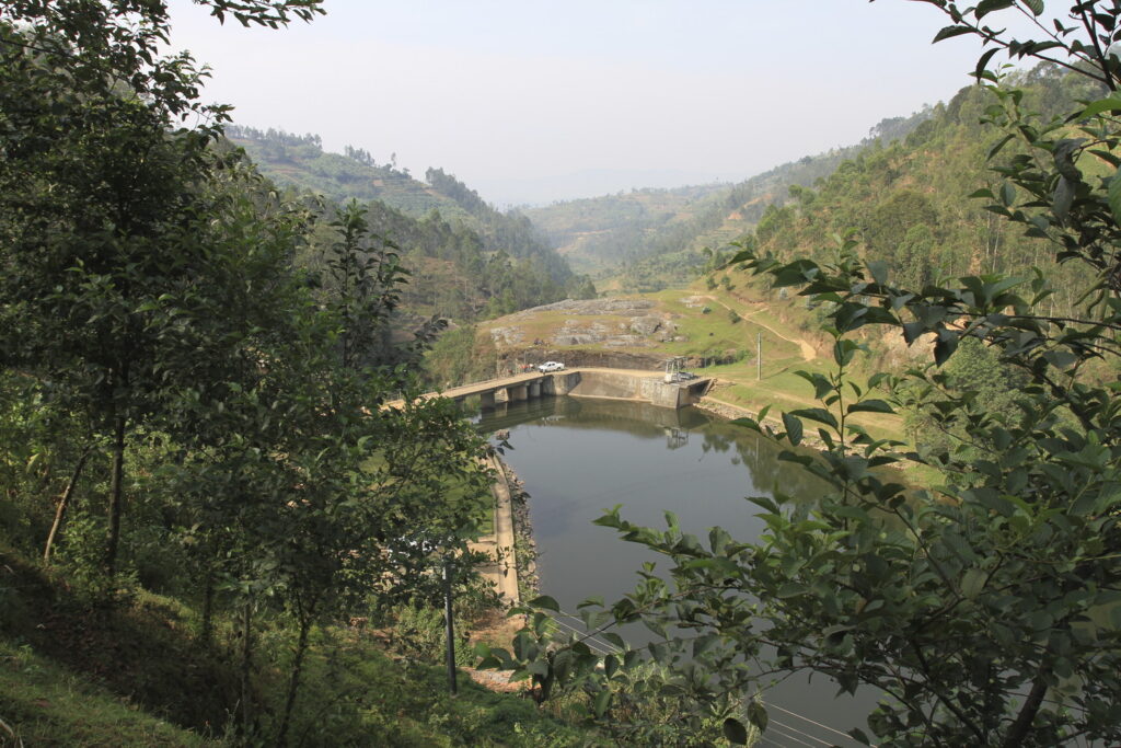 A small dam spans a river in a hilly, forested landscape with cars parked on the dam and surrounding lush greenery.