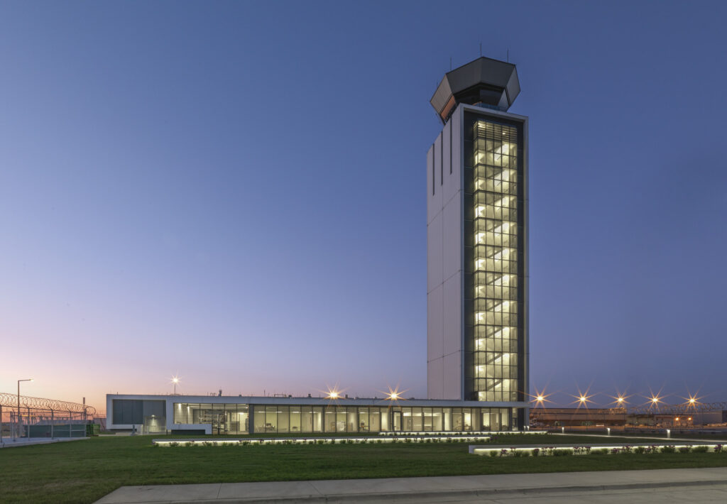 A tall, modern air traffic control tower stands illuminated at dusk, with a low building connected at its base. The surrounding area is flat with grass and pavement.