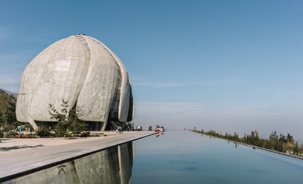 A lotus-shaped building with reflective glass exterior stands beside a long, rectangular reflecting pool under a clear blue sky, with trees and people in the distance.