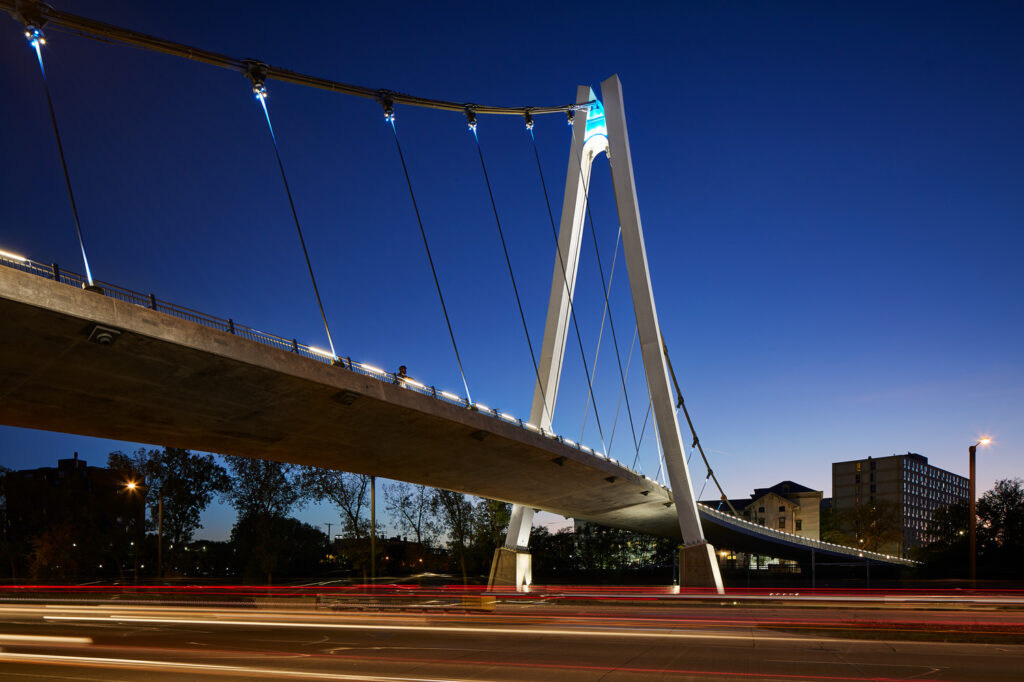 A modern cable-stayed bridge illuminated at night with vehicular light trails visible on the road below. The bridge features a tall, central pylon and a clean architectural design.