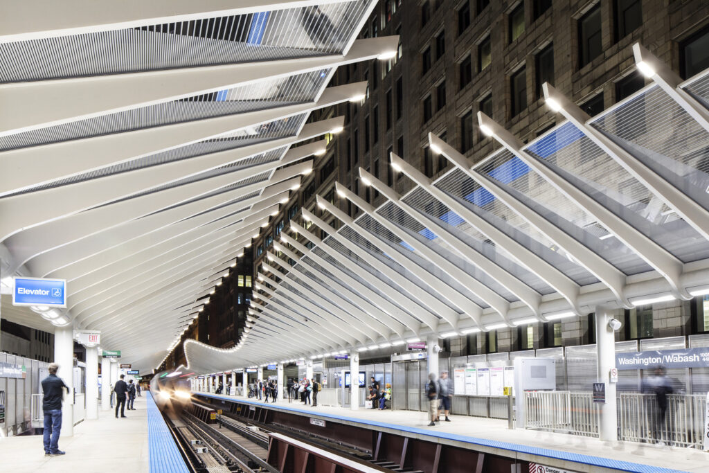 Modern train station with an open-air design and white ribbed roof structure. Passengers wait on the platform, and an approaching train is visible. The sign reads "Washington/Wabash.