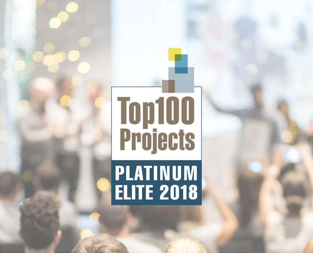 A large room with many people in focus on an award placard at the center that reads "Top 100 Projects Platinum Elite 2018.