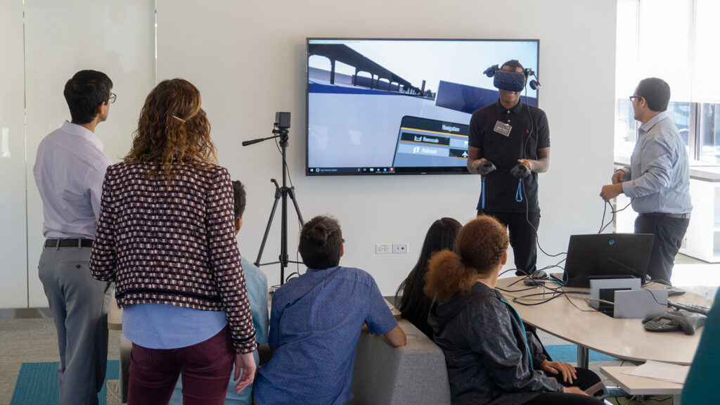 A group of people in an office setting watch a demonstration of virtual reality technology, with one person wearing a VR headset and holding controllers.