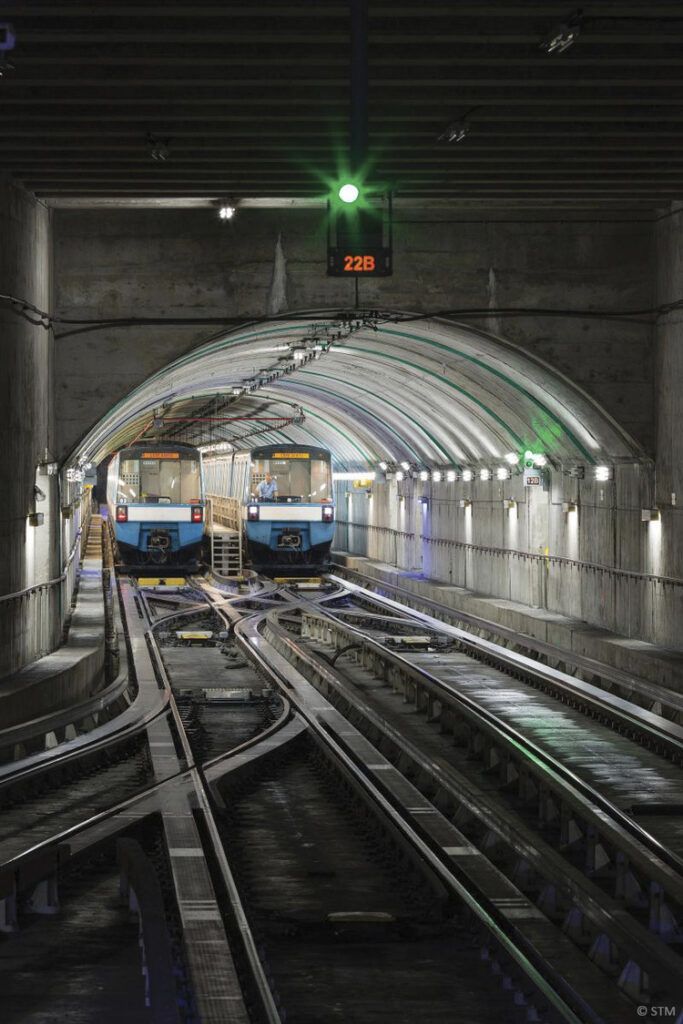 Two subway trains are stationed on parallel tracks in a dimly lit underground tunnel, illuminated by a green signal light overhead. The tunnel walls are lined with lights.