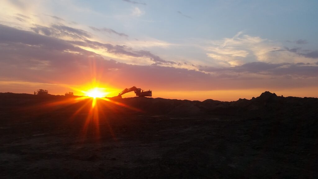 A silhouette of construction equipment against a sunset sky, with clouds and a mix of orange, yellow, and blue hues in the background.