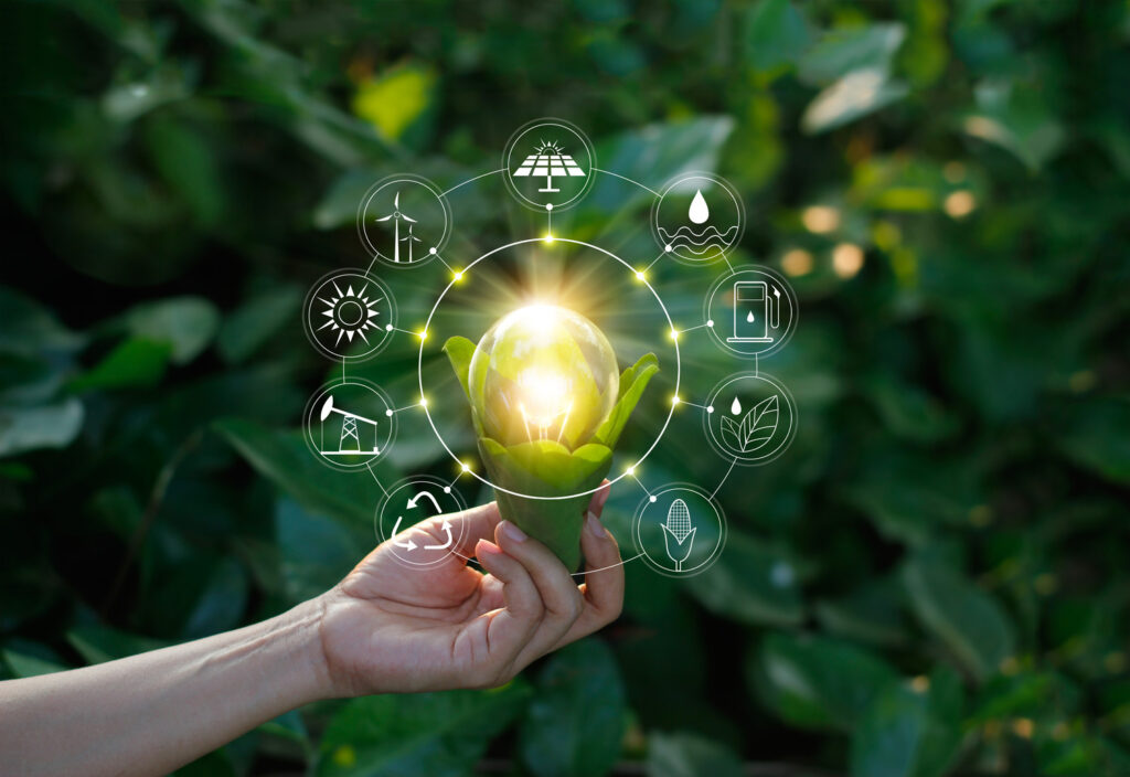 A hand holding a glowing light bulb encased in green leaves, surrounded by symbols of renewable energy sources such as wind, solar, and water, against a background of leafy greenery.
