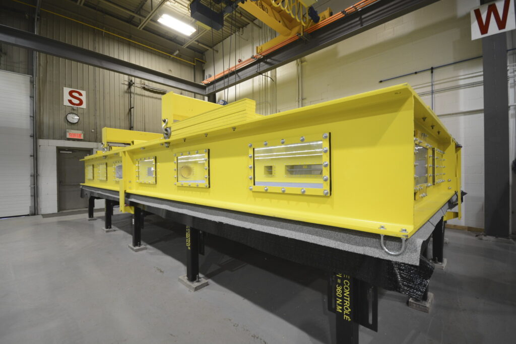 Large yellow industrial machinery piece with multiple inspection windows, mounted on black support beams in an indoor facility with concrete flooring and grey walls.