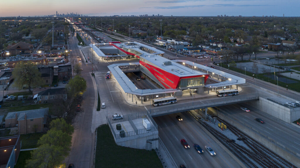 Aerial view of a modern transportation station with a red roof, multiple levels, buses, and trains. The surrounding urban area is visible, with a city skyline in the background under a cloudy sky.