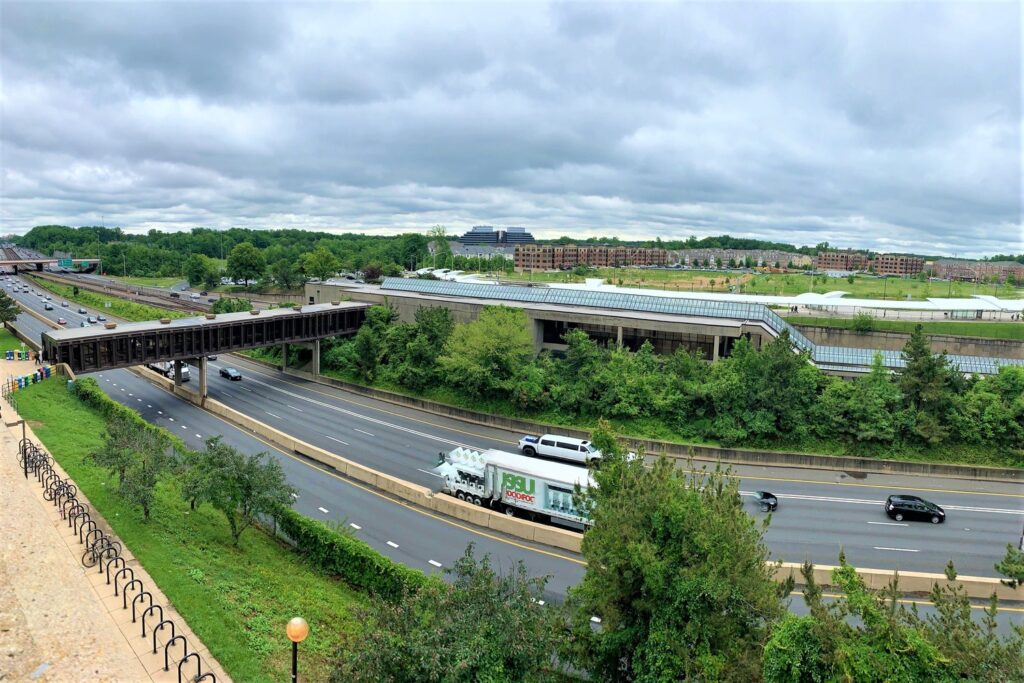 A wide-angle view of a highway with a few vehicles, bordered by greenery. An overpass crosses the highway, with buildings and cloudy skies in the background.